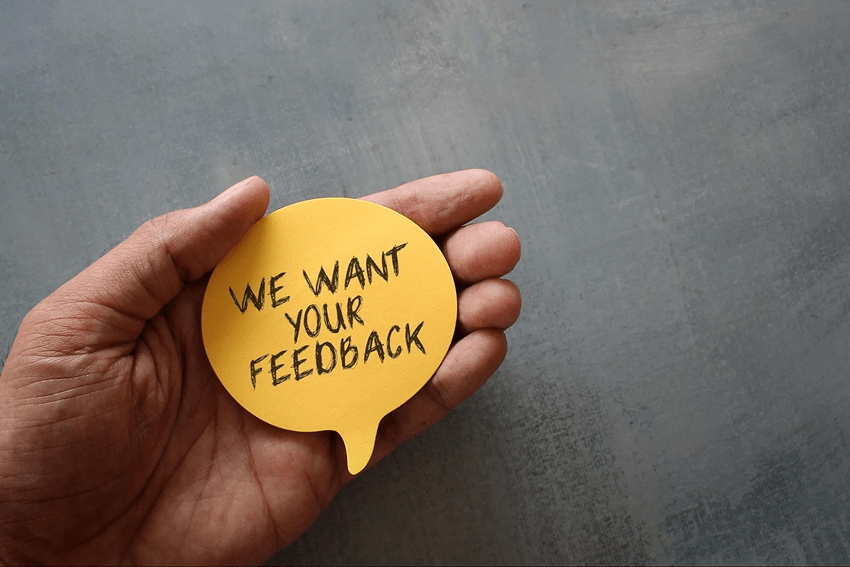 Close-up of a hand holding a thinking bobblehead with the message We want your feedback