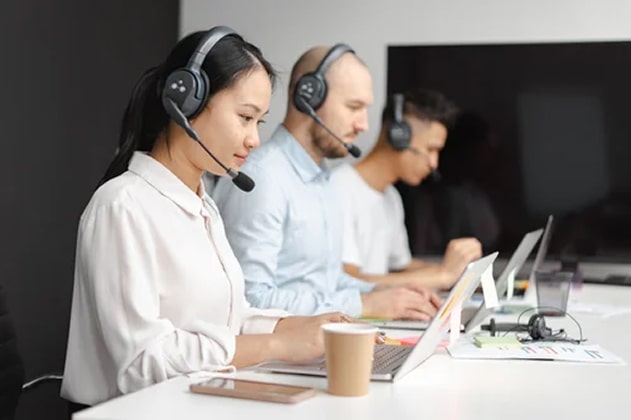 3 people sitting in front of laptops wearing headphones, working in customer service.