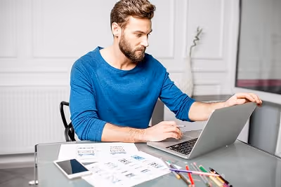 A man working on his laptop and holding a pencil while being surrounded by documents, pens, and his 