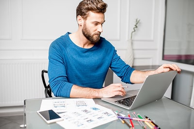 A man working on his laptop and holding a pencil while being surrounded by documents, pens, and his 