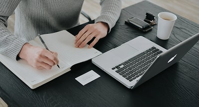 Someone in front of a laptop writing in a notebook. Photo by Vadim Bozhko on Unsplash