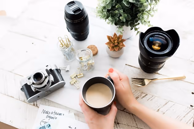 Image of a table, a camera and lens sitting on the table, a set of hands holding a mug