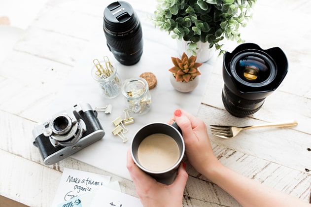 Image of a table, a camera and lens sitting on the table, a set of hands holding a mug