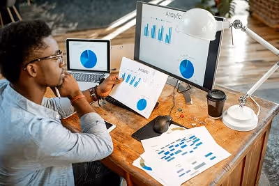 A man looking at statistics on a sheet of paper, a laptop and a monitor. A man looking at statistics on a sheet of paper, a laptop and a monitor.