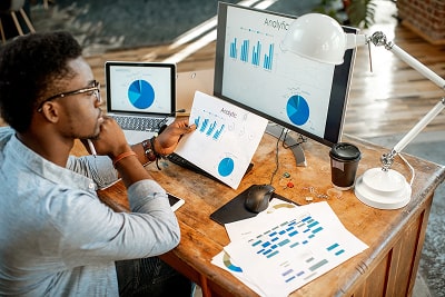 A man looking at statistics on a sheet of paper, a laptop and a monitor.