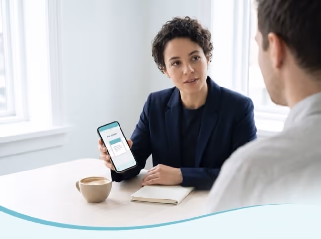A salesperson holds up a phone showing the one deck page that answers the buyer's question, while the buyer listens, seen from behind. A salesperson holds up a phone showing the one deck page that answers the buyer's question, while the buyer listens, seen from behind.