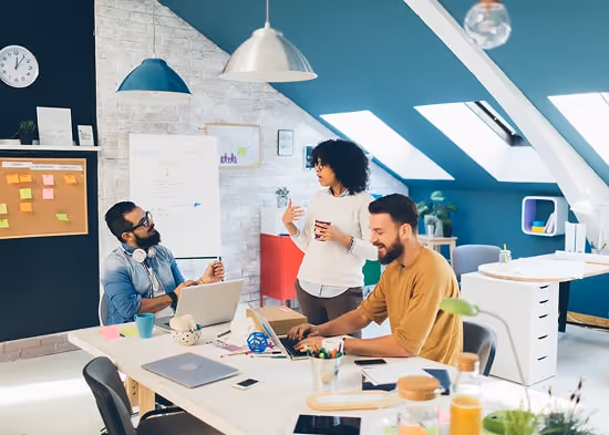 In a loft office, three people are having a meeting. Two are sitting around a table with their compu