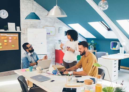 In a loft office, three people are having a meeting. Two are sitting around a table with their compu