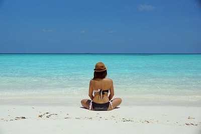 A woman waring a bikini and sitting with her back to the camera on a beach watching the clear blue w