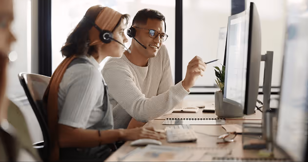 Two colleagues wearing headsets, discussing something while looking at a computer screen. Two colleagues wearing headsets, discussing something while looking at a computer screen.