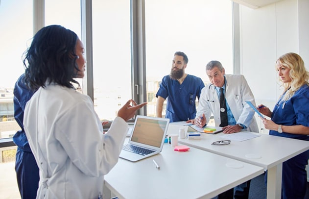Doctors in lab coats discuss medical records at a table with medication bottles and a computer.