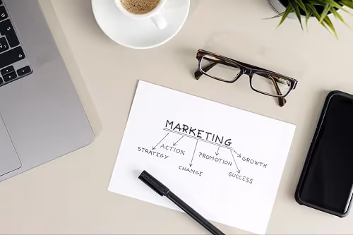 A desk with a laptop, phone, glasses, and a paper with a marketing strategy outlined on it. A desk with a laptop, phone, glasses, and a paper with a marketing strategy outlined on it.